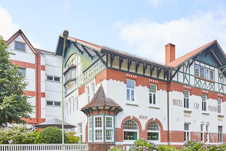 Aussenaufnahme in Schrägansicht des pittoresken, im Jugendstil 1908 erbauten Hotel Cap Polonio mit weißer Fassade, grünem filigranem Fachwerk, roten Ziegel-Schmuckelementen, großen Sprossen-Fenstern und einem runden Erker an der linken Hausecke im Erdgeschoss. Auf der Fassade befindet sich der rote Serifen-Schriftzug "Hotel Cap Polonio" und "Rolin". Linksseitig angeschnitten ist der moderne und schlichte Hotelanbau sichtbar.