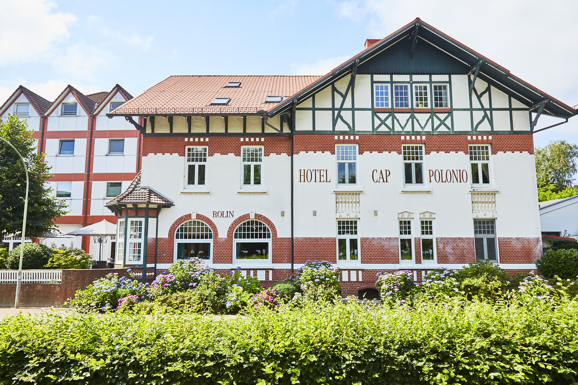 Aussenaufnahme in Frontalansicht des pittoresken, im Jugendstil 1908 erbauten Hotel Cap Polonio mit weißer Fassade, grünem filigranem Fachwerk, roten Ziegel-Schmuckelementen, großen Sprossen-Fenstern und einem runden Erker an der linken Hausecke im Erdgeschoss. Auf der Fassade befindet sich der rote Serifen-Schriftzug "Hotel Cap Polonio" und "Rolin". Linksseitig angeschnitten ist der moderne und schlichte Hotelanbau sichtbar.