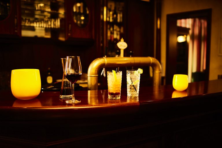 Stimmungsvolle Bar "Deck 48" mit dezentem warmem Licht: Auf der polierten Holztheke stehen zwei Cocktails, ein Glas Rotwein und ein Glas Wasser, eingerahmt von zwei leuchtenden gelben Windlichtern. Im Hintergrund ist ein klassischer Bierzapfhahn und ein Regal mit Gläsern zu sehen.