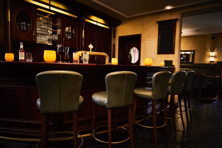 Elegante Hotelbar "Deck 48" dezent beleuchtet mit dunkler Holzverkleidung, Messingzapfanlage und sechs grünen Barhockern mit Lederpolsterung. Auf der Theke stehen mehrere leuchtend gelbe Windlichter, die dem Raum eine warme Atmosphäre verleihen. Im Hintergrund ein Spiegel, Wandlampen und eine Tafel mit Tagesangebot.