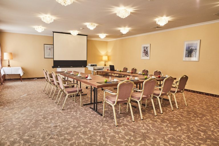Tagungsraum im Hotel Cap Polonio mit U-förmigen Holztischen, Stühlen, einem Projektor, einer Projektionsleinwand und Gedecken wie Notizblöcken, Tassen, Gläsern, Stiften und Äpfeln. Der Raum ist mit Lampen und Wandkunst warm beleuchtet.