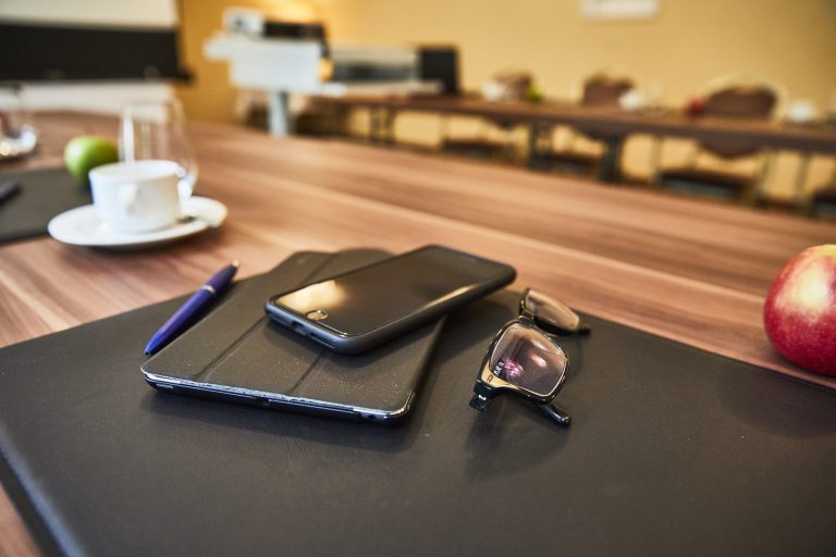 Nahaufnahme im Tagungsraum des Hotel Cap Polonio eines Konferenztisches mit einem schwarzen Smartphone, einem Tablet mit Hülle, einer Brille und einem Kugelschreiber auf einer schwarzen Schreibunterlage. Im Hintergrund stehen eine Tasse mit Untertasse, ein Glas Wasser, ein grüner Apfel und ein roter Apfel auf dem langen Tisch.