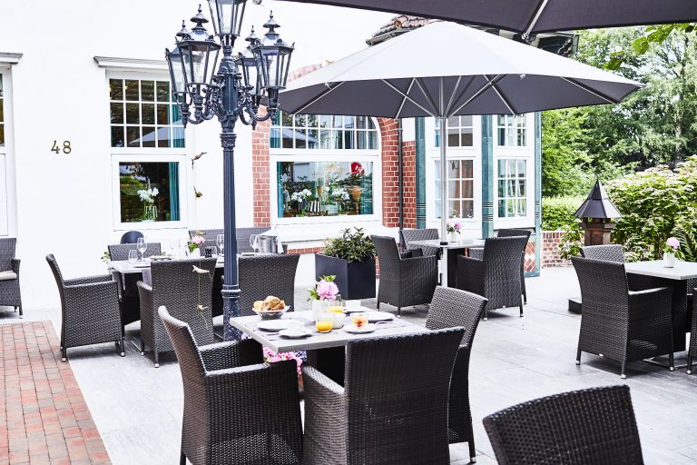 Die Restaurant-Terrasse im Freien mit Korbtischen und -stühlen, Frühstücksgerichten und Getränken auf den Tischen, einem großen Sonnenschirm, einem dekorativen Laternenpfahl und Grün im Hintergrund schaffen eine einladende Atmosphäre.