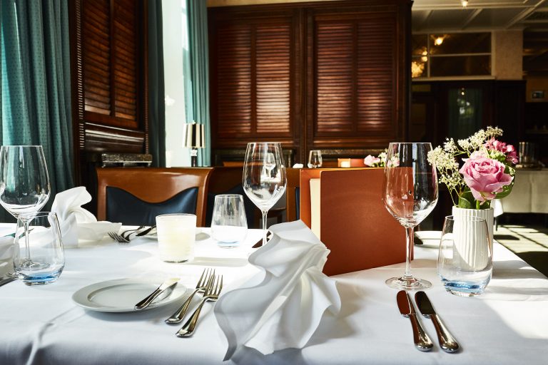 Ein gedeckter Tisch im Restaurant Rolin mit Weingläsern, gefalteten weißen Servietten, Besteck, einer Kerze, einem Teller, einer Speisekarte und einer Vase mit rosa und weißen Blumen.