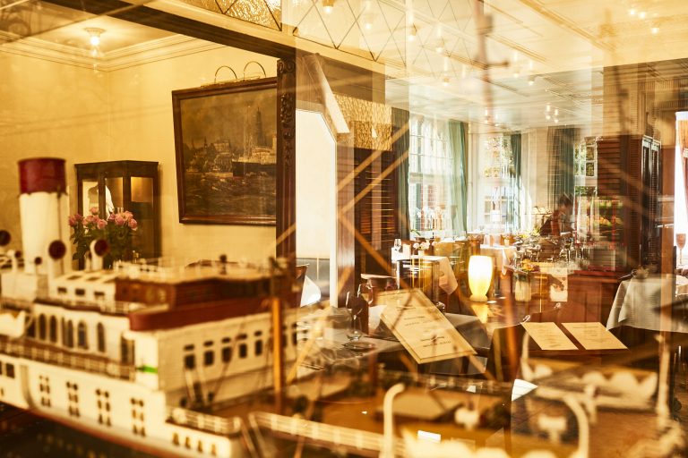 Durch eine Glasvitrine, die ein detailliertes Schiffsmodell der Cap Polonio zeigt, blickt man auf das warm beleuchtete Restaurant-Interieur mit elegant gedeckten Tischen. Ein Gemälde der Cap Polonio im Hamburger Hafen schmückt die Wand.