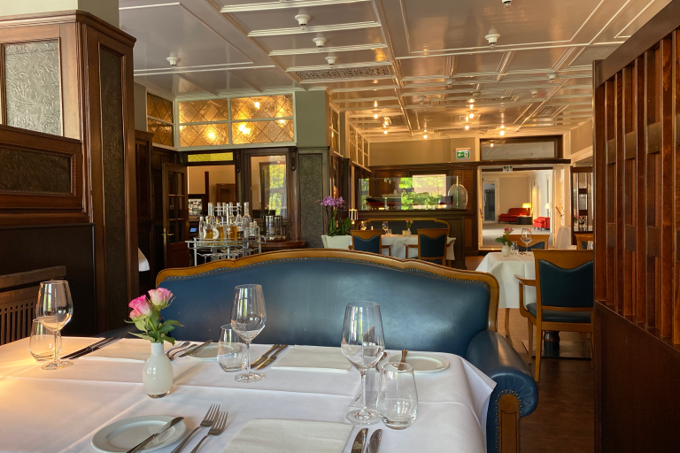 Ein gedeckter Tisch im Restaurant Rolin mit weißem Tischtuch, Gläsern, Besteck und rosa Rosen neben einer blau gepolsterten eleganten Bank. Die Wände sind mit dunklem Holz vertäfelt, die Decke mit weißlackiertem Holz. Die Original-Vertäfelung und Teile der Einrichtung stammen aus dem Passagierschiff Cap Polonio. Im Hintergrund sind weitere gedeckte Tische und eine elegante Bestuhlung zu erkennen.