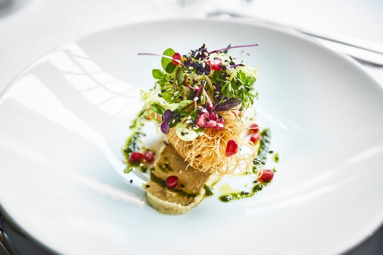 Ein Gourmet-Gericht im Restaurant Rolin mit einer knusprigen Vorspeise aus regionalen Produkten und frischen Kräutern, belegt mit Granatapfelkernen, serviert auf einer cremigen Sauce mit Kräuteröltupfern.