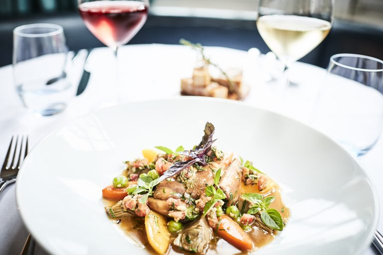 Ein weißer Teller mit einem Gourmet-Gericht aus Fisch, Gemüse und Kräutern steht auf einem gedeckten Tisch im Restaurant Rolin, in dem Speisen aus regionalen Produkten und frischen Kräutern serviert werden, begleitet von Gläsern mit Weiß- und Rotwein im Hintergrund.