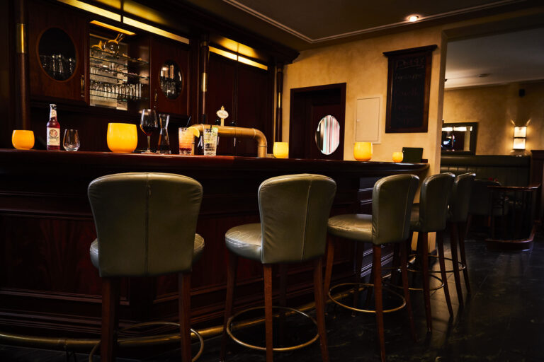 The dimly lit bar ‘Deck 48’ with five leather-covered empty stools at a polished wooden counter on which several glasses, two cocktails, a bottle of beer and orange candles are placed. Behind the bar, a dark wood and glass shelf can be seen for storing more glasses.