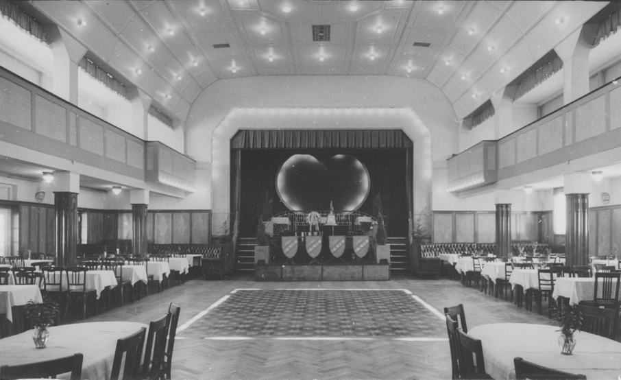 A black-and-white photograph from the early days of the hotel shows the historic ballroom of the Hotel Cap Polonio with a dance floor and tables set under a gallery on the left and right. The stage curtain is open, and heart-shaped lighting is located on the back wall.