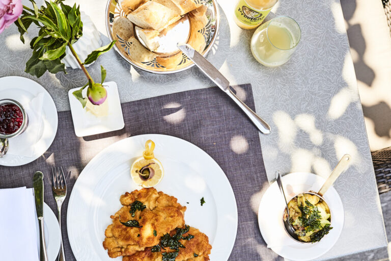 A table set outdoors with a plate of Wiener schnitzel garnished with lemon and parsley. Next to it is a glass of lemonade, a small pot of fried potatoes, cranberries, a bread basket and a pink flower in a vase.