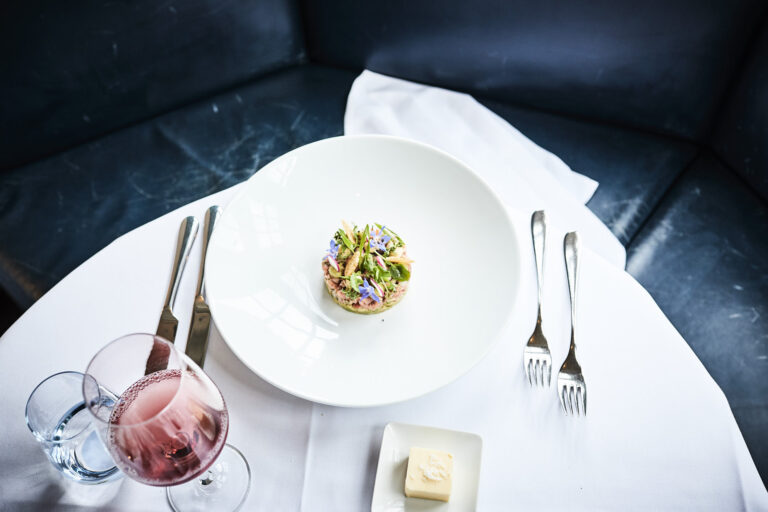 A small, colourful gourmet plate made from regional products and fresh herbs stands on a large white plate in the middle of a white tablecloth. Cutlery, a glass of rosé, water and butter complete the table setting.
