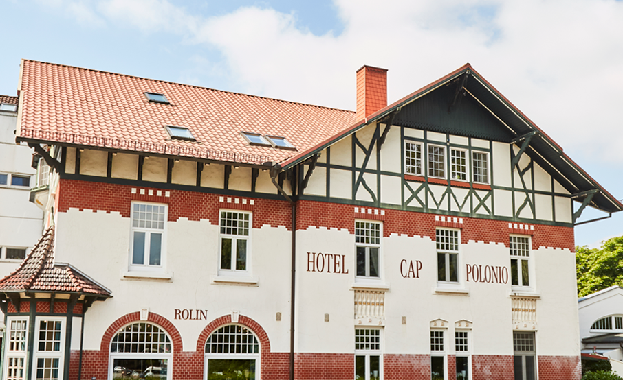 Exterior view in oblique angle of the picturesque Hotel Cap Polonio, built in Art Nouveau style in 1908, with a white façade, green filigree half-timbering, red decorative brick elements, large lattice windows and a round bay window on the left corner of the ground floor. The red serif lettering ‘Hotel Cap Polonio’ and ‘Rolin’ can be seen on the façade. The modern and simple hotel extension is visible on the left-hand side.
