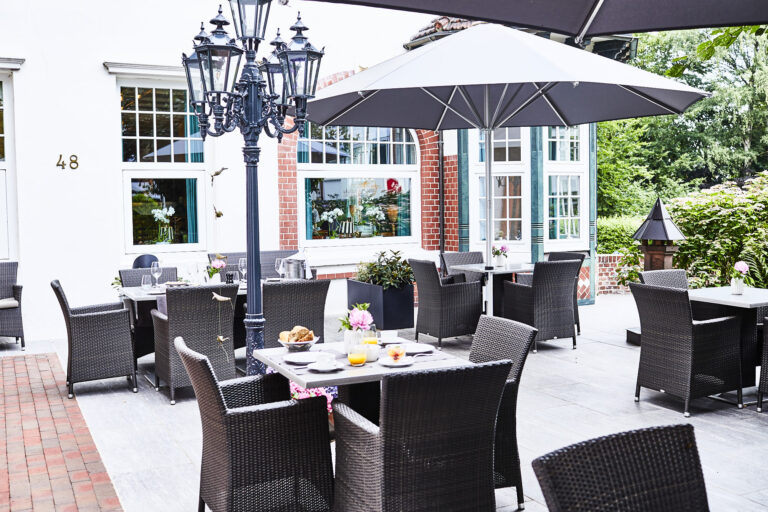 The outdoor restaurant terrace with wicker tables and chairs, breakfast dishes and drinks on the tables, a large parasol, a decorative lamppost and greenery in the background create an inviting atmosphere.