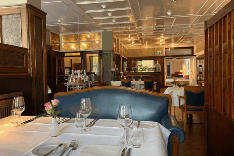 A table set in the Rolin restaurant with a white tablecloth, glasses, cutlery and pink roses next to an elegant blue upholstered bench.