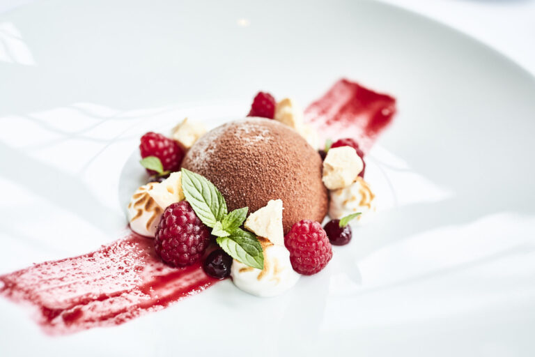 A round chocolate mousse dessert on a white plate, surrounded by raspberries, mint leaves, meringue balls and a red berry sauce.
