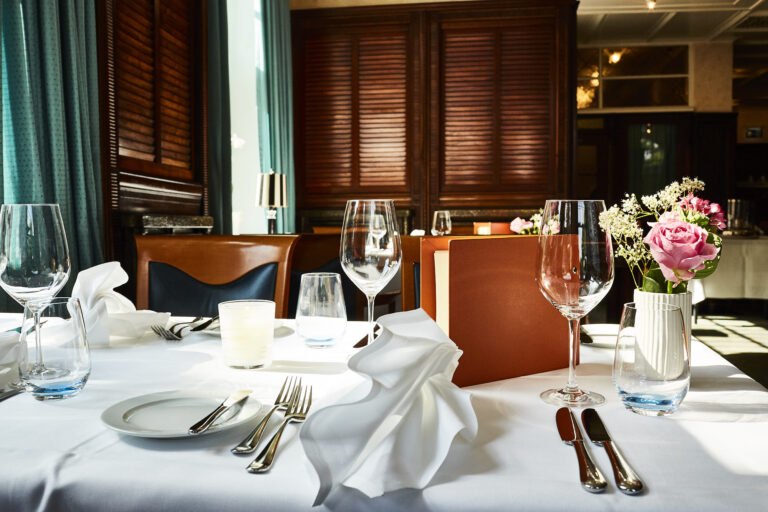 A table set in the Rolin restaurant with wine glasses, folded white napkins, cutlery, a candle, a plate, a menu and a vase with pink and white flowers.