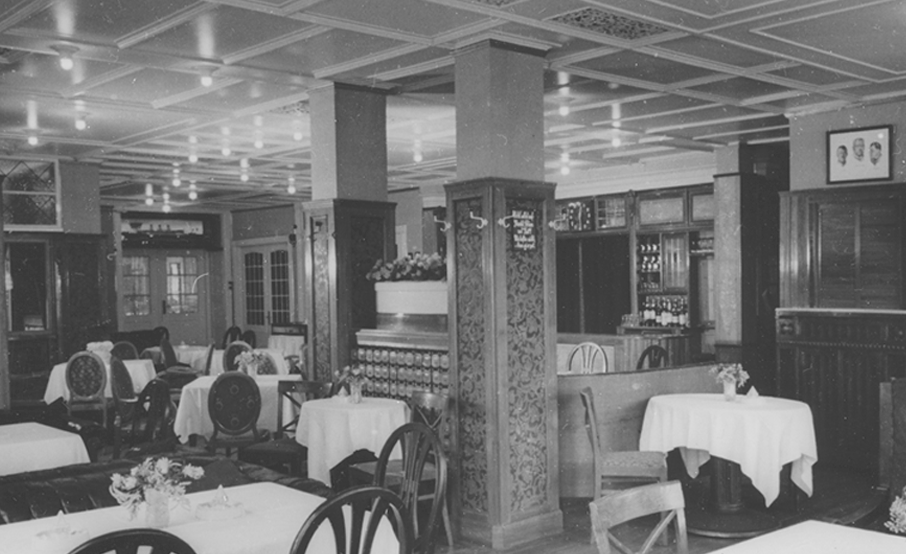 Black-and-white photograph of the Rolin Restaurant from the hotel's early days. Tables set with elegant chairs and white tablecloths. The walls are panelled in dark wood and the ceiling is painted white. The original panelling comes from the dining room of the passenger steamship Cap Polonio. Other furnishings from the Cap Polonio can also be seen, such as a large wooden cabinet and decorative elements.