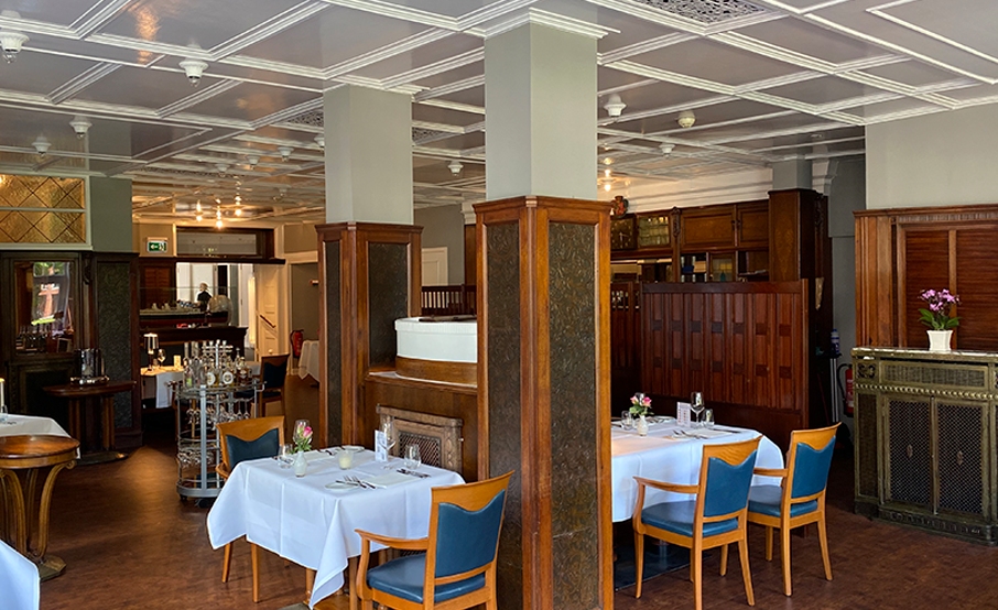 Two tables set with elegant tableware in the Rolin restaurant with white tablecloths, glasses, cutlery and pink roses. The walls are panelled in dark wood and the ceiling is painted white. The original panelling comes from the passenger ship Cap Polonio. In the background, you can see more tables and a large wooden cabinet, which also belonged to the Cap Polonio.