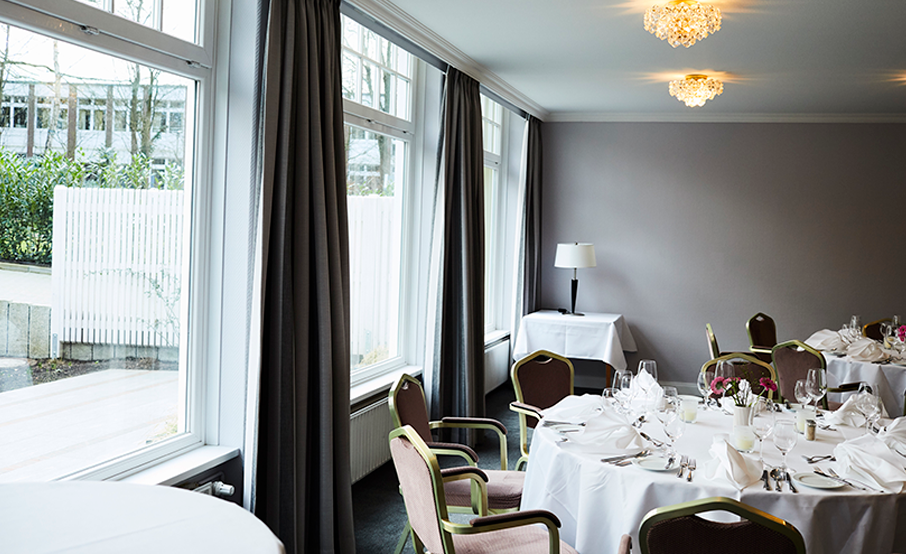 Room prepared for a conference at the Hotel Cap Polonio with large windows, round tables with white tablecloths, glasses and napkins. Brown chairs surround the tables.