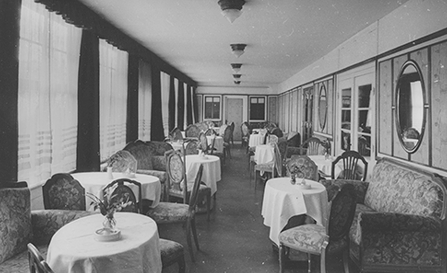 Black-and-white photograph of a narrow, elongated room in the Rolin restaurant from the early days of the hotel. Tables covered with white tablecloths, elegant chairs and upholstered benches. Oval mirrors hang on one wall. The curtains on the windows of the opposite wall are drawn.
